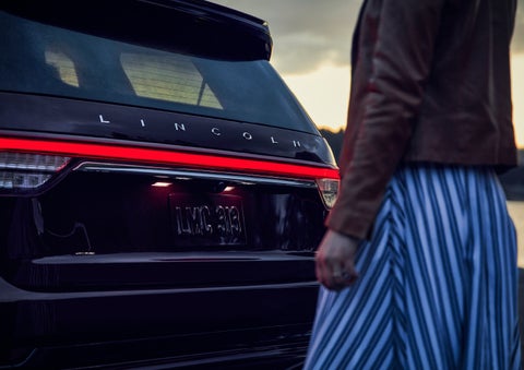 A person is shown near the rear of a 2024 Lincoln Aviator® SUV as the Lincoln Embrace illuminates the rear lights | Koons Lincoln of Bethesda in Silver Spring MD